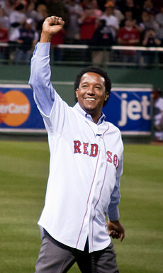 A man with dark curly hair stands on a grass field. He is wearing a white baseball jersey with "RED SOX" across the chest in red type.