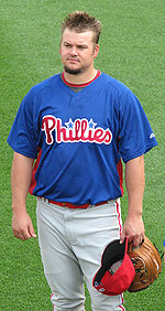 A man with spiked hair and a goatee wearing a blue baseball jersey with "Phillies" in red across the front and white baseball pants with red pinstripes stands on a baseball field holding a tan baseball glove and a red baseball cap in his left hand.