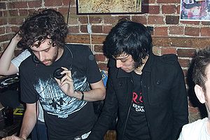 A color photograph of two members of the group Justice standing in front of a brick wall