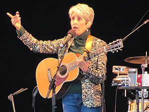 Baez holds guitar, in blue jeans, brown mock turtleneck, patterned jacket, black backdrop, talking and gesturing