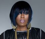 Missy Elliott posing on a grey background wearing a black shirt and gold jewelry.