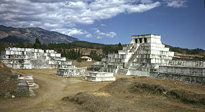 A cluster of squat white step pyramids, the tallest of them topped by a shrine with three doorways. In the background is a low mountain ridge.