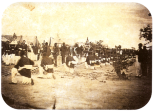 An old photograph showing a procession passing between lines of soldiers with tents in the background