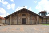 A church and bell-tower in frontal view. The whitish facade is decorated with motifs painted in orange. A wooden cross is positioned at the top of the roof.