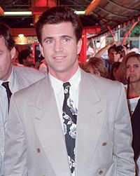 A man in a grey suit. Behind him is a small crowd of people.