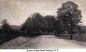 A highway flanked on both sides by fence and trees proceeds through mostly open land.