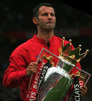 A head-and-torso photograph of a man with dark hair wearing a red tracksuit top. He is holding a large silver trophy with a gold crown on top. The trophy is decorated with one red ribbon and one black ribbon attached to each handle.