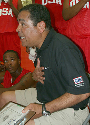 A man wearing a gray shirt with a clipboard in hand surrounded by basketball players all wearing red jerseys with "USA" on the chest.