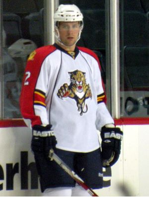 A Caucasian ice hockey player standing relaxed on the ice. He wears a white, visored helmet and a white and orange jersey. He holds his stick on the ice lightly with one hand.