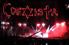 A view of the stage being filmed, with "COEXISTA" spelled out on the video screen, using an Islmaic crescent as a "C", a Star of David as an "X", and a Christian cross at a "T". A camera in the middle shows the concert being filmed.