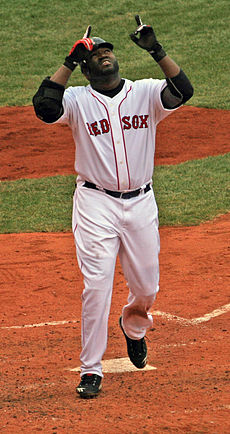 A dark-skinned man in a black baseball jersey and gray pants takes a right handed baseball swing with a crowd in the background, several people wearing red.