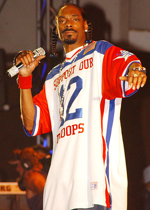 An African American male with his black hair in braids. The male is holding a microphone with his right hand. The male is wearing a shirt that has the colors red, blue and white in the form of writing, numbers, and stripes.