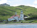 alt=Photograph of the Church of the Holy Ascension on a sunny day, with red roofs, green onion domes, and a small churchyard.