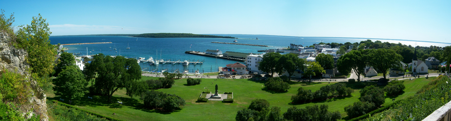 A panoramic view of a park. To the left is a rocky cliff. A wide green space, named Marquette Park, can be seen in the foreground. Ships are docked in a harbor in the background, and another island is visible in the distance.