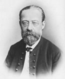 Portrait of balding, bearded, bespectacled middle-aged man with solemn expression, wearing a bow tie and high-buttoned jacket