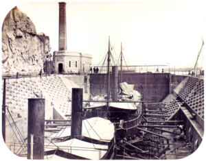 A photograph with two steamships resting in a dewatered drydock with a building housing the engine for operating the lock's gates in the background