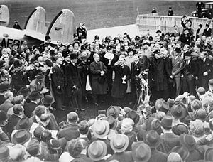 A large crowd on an airfield; British Prime Minister Neville Chamberlain presents an assurance from German Chancellor Adolf Hitler.