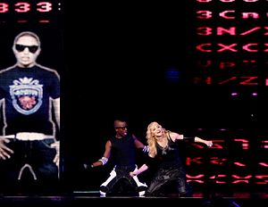 A blond woman sitting on a stage on her knees, flanked by an African-American man on her right, in the same position. They are both dressed in black pants and vests, with broad rubber bands on their hand. To their right, a vertical rectangular screen shows a man in a black T-shirt standing with his hands on his waist and wearing sunglasses.