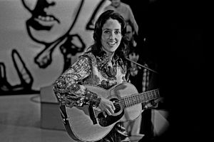 Baez is seated, wearing long-sleeve paisley shirt, smiling