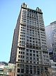 Ground-level view of a 30-story building with a tan exterior and a square cross-section; spires sit atop the roof of the building at each corner