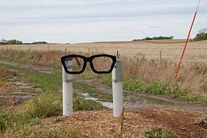 A sculpture of sort, consisting of two white posts holding a black spectacles frame in Buddy Holly's characteristic style