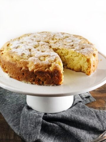 The rustic Italian apple cake on a cake stand with a slice cut out of it.