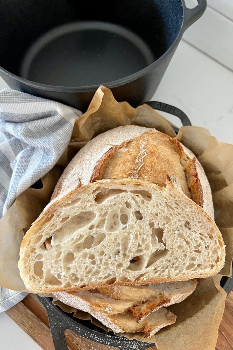 Slice of Dutch Oven Sourdough Bread sitting on a loaf that has been baked in a Lodge Cast Iron Dutch Oven. The Empty Dutch Oven can be seen in the background of the photo.