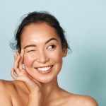 Skin care. Woman with beauty face touching healthy facial skin portrait. Beautiful smiling asian girl model with natural makeup touching glowing hydrated skin on blue background closeup