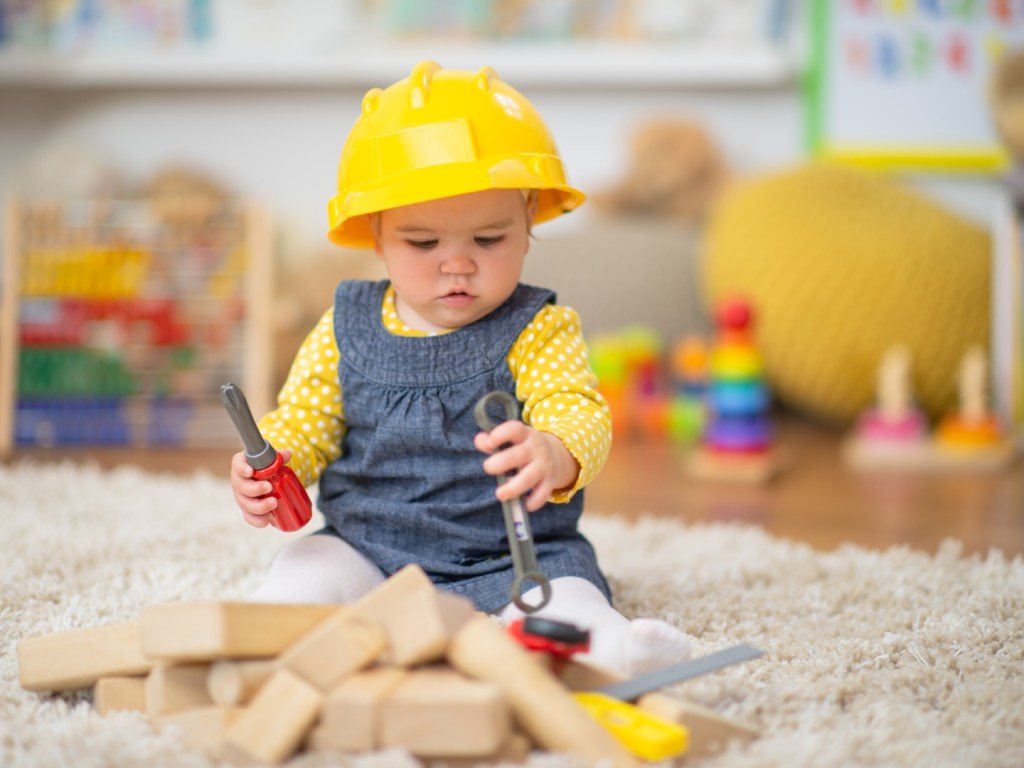 An adorable little girl has a yellow construction hat on her head and is holding some tools. She is sitting down on a carpet and there are wooden blocks in front of her.