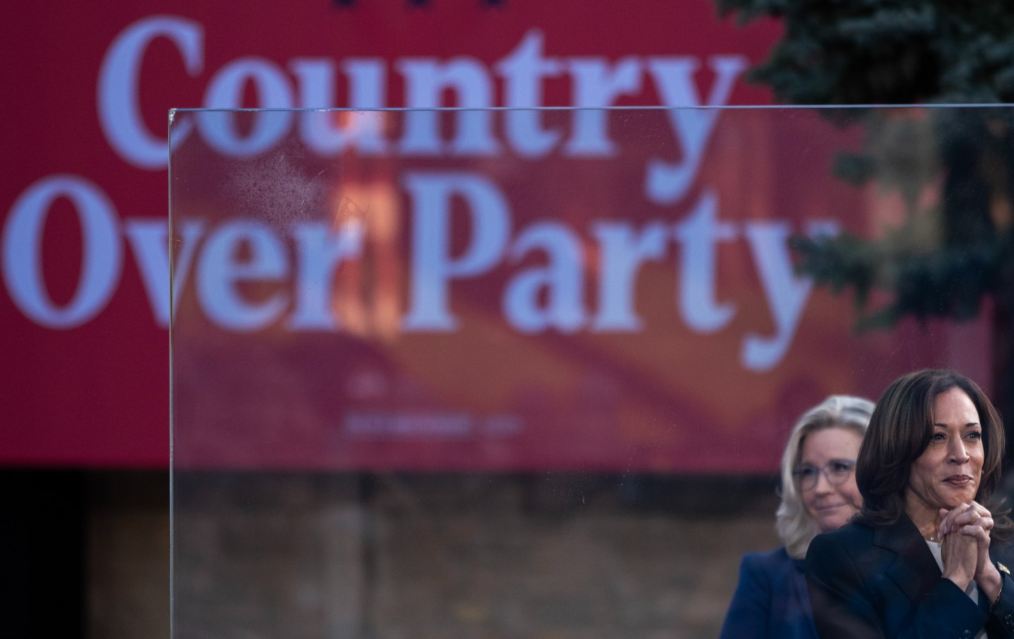 Kamala Harris, with Liz Cheney behind her, smiles and clutches her hand. A banner reads Country Over Party behind.