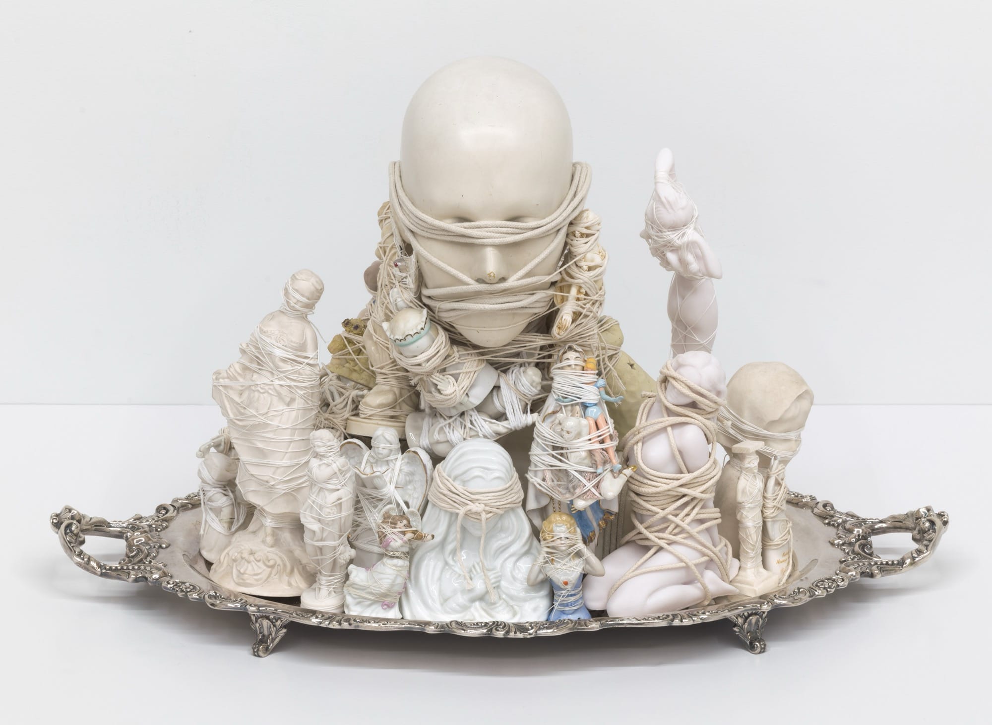 a sculpture made from found objects and figurines tied together with string atop a silver serving platter