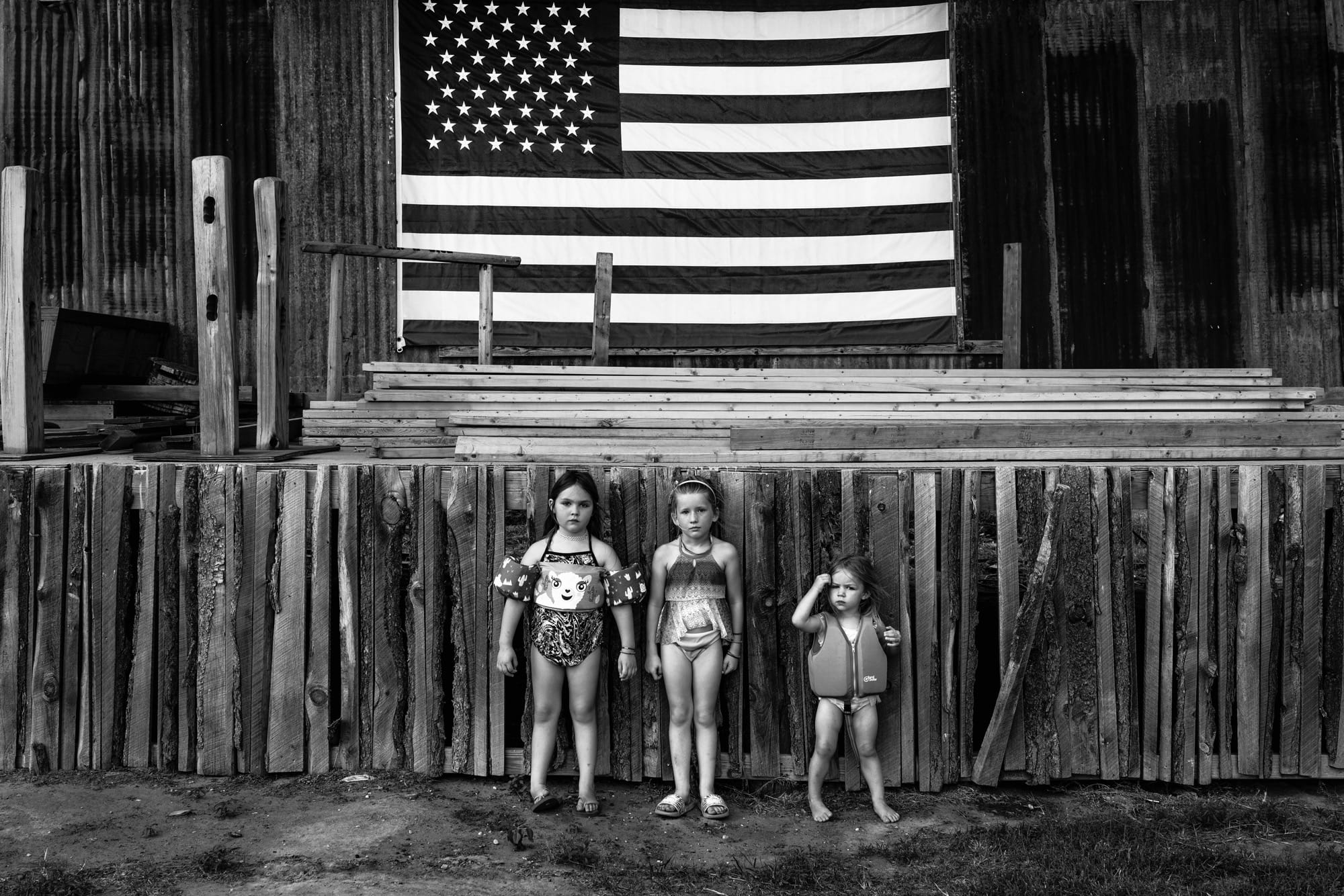 a black and white photo of three young girls in bathing suits standing in front of a wodoen stage with an american flag in background