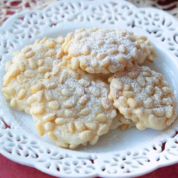 pignoli cookies served on a decorative white serving dish on a maroon tablecloth with white lace placed on top of the tablecloth with another plate of pignoli cookies and tea in the background