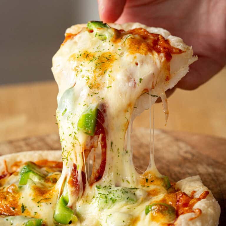 A hand removes a slice of mochi cheese pizza with green capsicum.