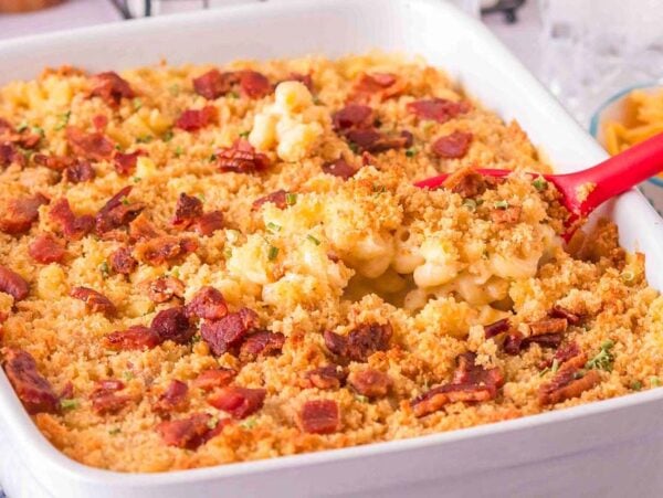 A loaded mac and cheese dish featuring baked macaroni drenched in creamy cheese, topped with savory breadcrumbs and crispy bacon bits, all nestled in a rectangular white casserole dish with a bright red serving spoon.