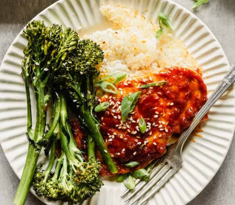 chicken breast coated in a red sauce plated over rice with broccolini in an off-white bowl