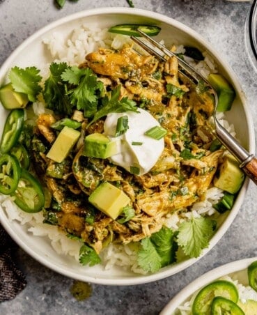 Shredded salsa verde chicken served over rice in a shallow white bowl topped with avocado, cilantro and jalapenos.